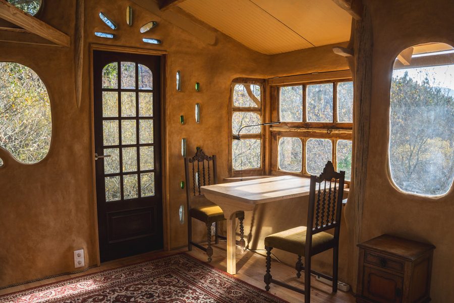 Inside the Clayhouse: earthen walls, a glazed door, glass bottles set in the clay and a table at the window