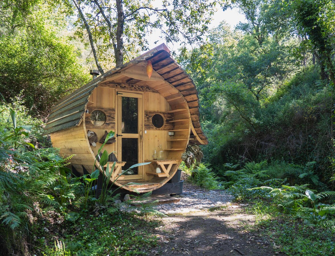 The barrel sauna with round windows either side of a glazed door, a curved shingled roof and a chimney, among ferns and oaks