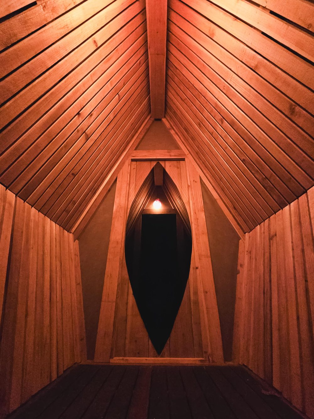 Inside the shrine at night: boarded walls and a raking ceiling of slats converging on the ridge, with the almond-shaped opening at the far end