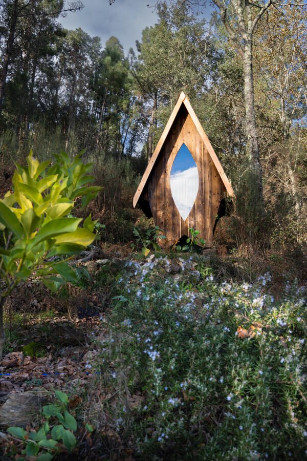 The shrine on a wooded hillside: a tall A-frame of weathered boards with an almond-shaped window catching the sky, rosemary in flower in front