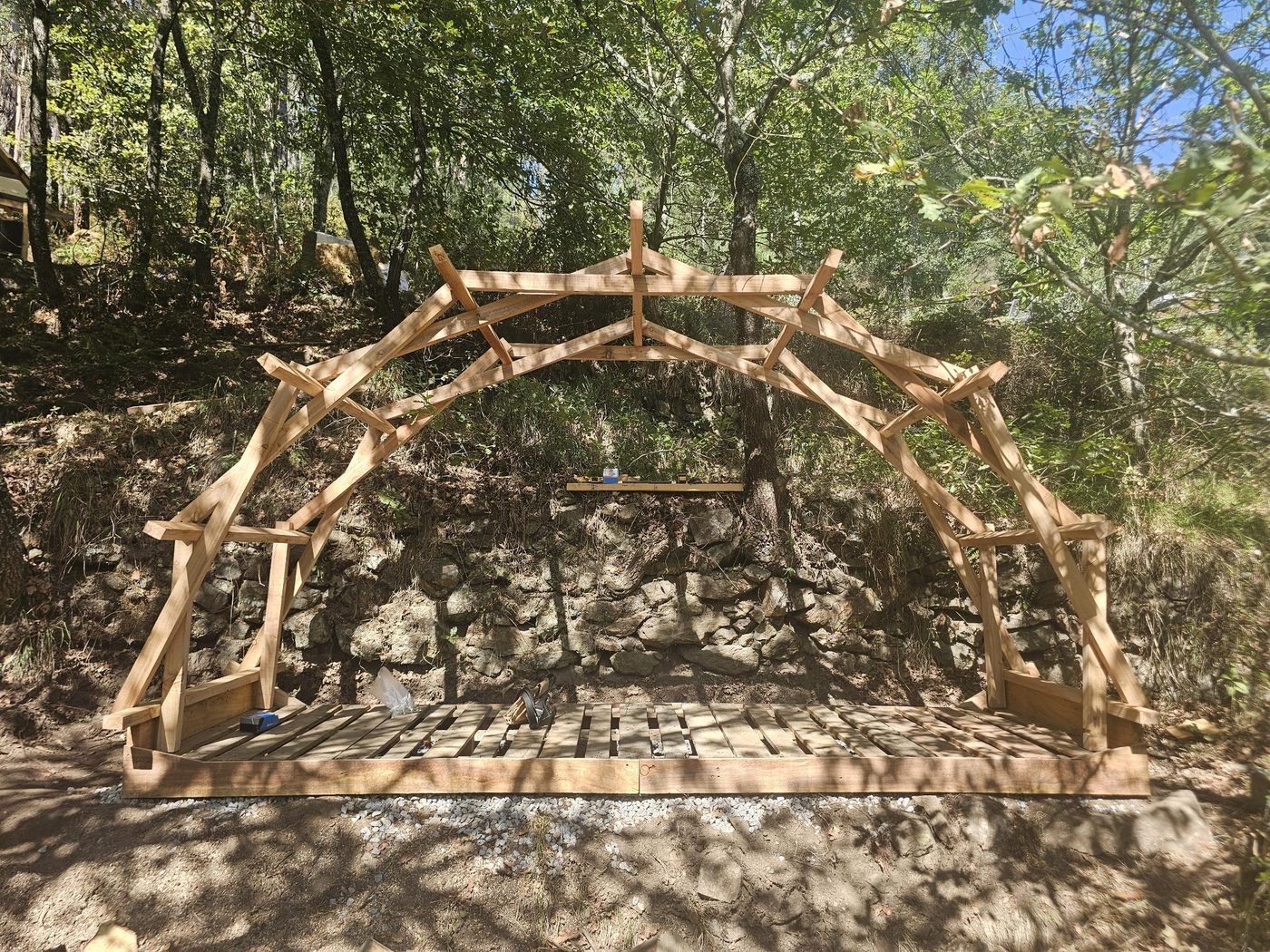 The bare reciprocal arch frame standing on its pallet deck in front of a dry stone wall